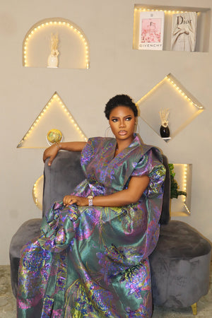 Woman in a teal Boubou dress sitting on a gray chair with decorative wall lights and framed pictures in the background.